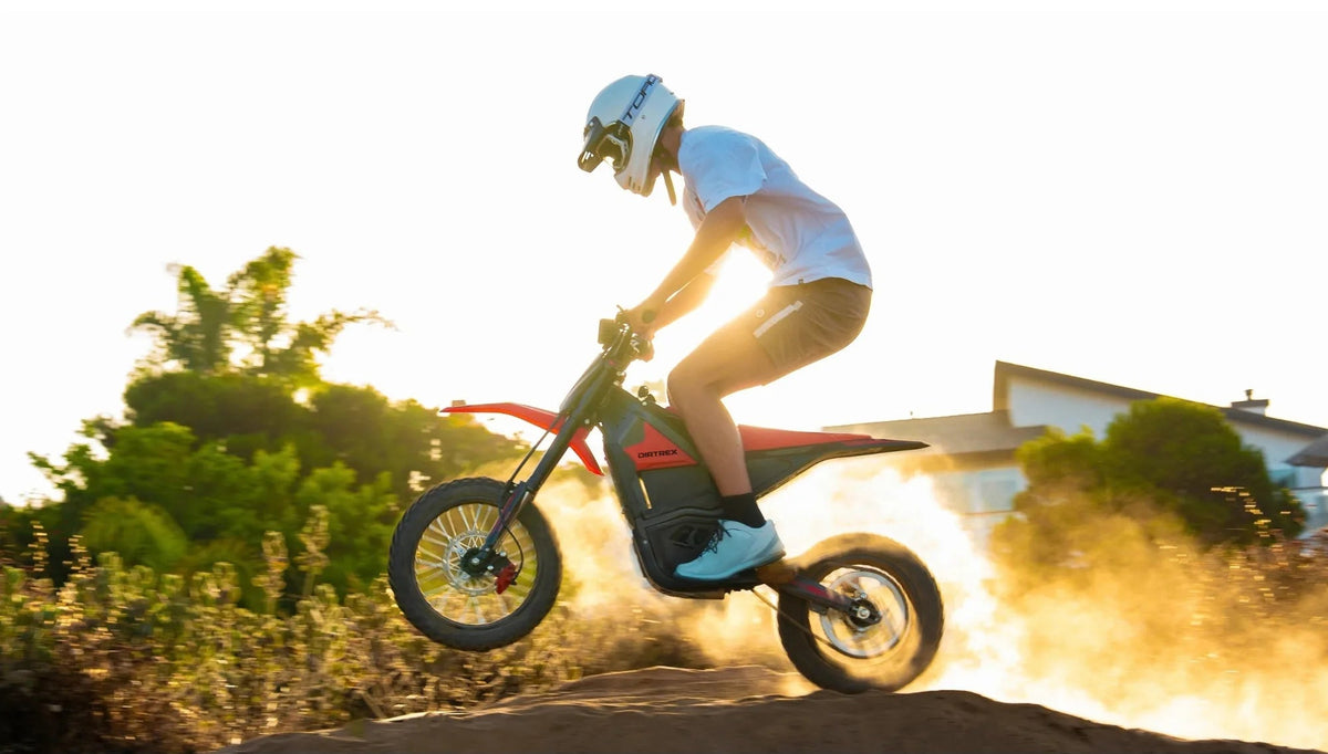 A person riding a red and black electric dirt bike on a dirt mound at sunset, with the rider wearing a white helmet, white t-shirt, and light blue sneakers kicking up dust in an outdoor setting.