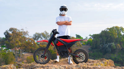 A person in a white t-shirt, black shorts, white sneakers, and a white helmet with goggles stands with arms crossed next to a red and black electric Dirtrex dirt bike on a rocky outdoor setting with trees in the background.